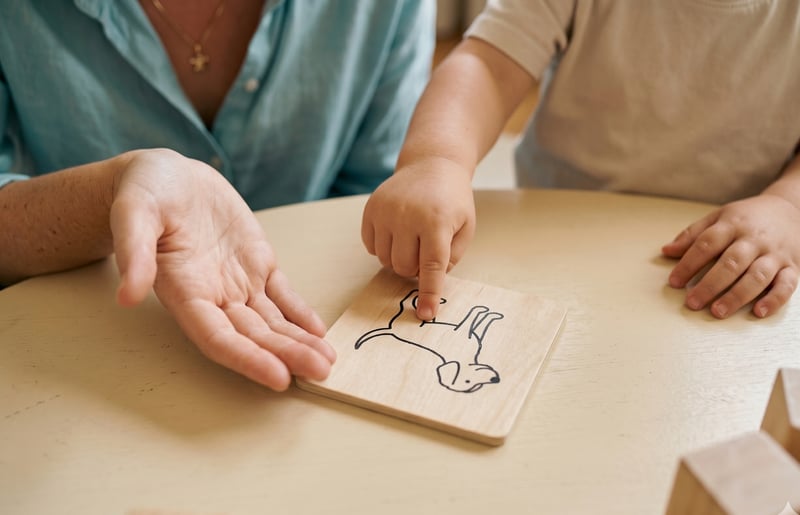 A speech pathologist and a young child seated at a low table, their hands together over a wooden picture card. The child's finger rests on a line drawing of a dog.